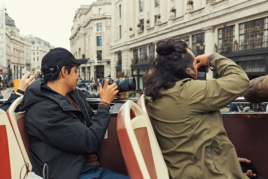 Travelers on an open top bus take photos of bustling city streets and architecture.